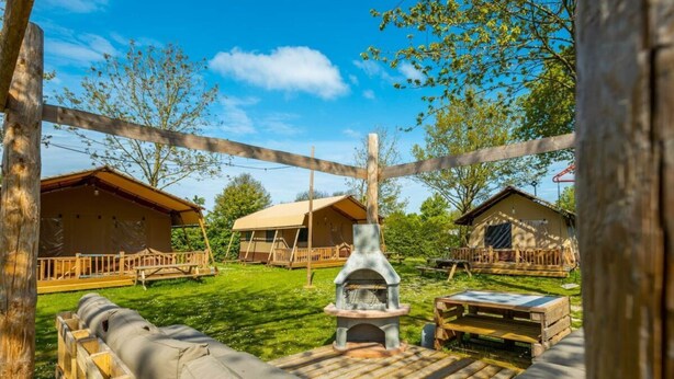 A rustic outdoor seating area with a stone fireplace, wooden deck, and several cabins with wooden railings. - Holiday Home in Noord-scharwoude With Wi-fi (Noord-Scharwoude)