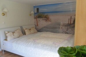 Ein Schlafzimmer mit Strandgemälde, einem weißen Bett mit flauschiger weißer Bettdecke und zwei Wandleuchten.