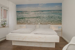 Ein Schlafzimmer mit großem Meereswandbild, einem Bett mit gestreifter Bettwäsche und einem weißen Stuhl.