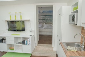 Eine kompakte Küche mit integriertem Fernseher, einer kleinen Spüle und Blick auf einen Strand durch eine offene Tür.