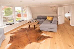Ein modernes Wohnzimmer mit einem grauen Ecksofa, einem Couchtisch aus Holz, einem Kuhfellteppich und großen Fenstern.