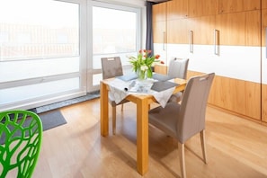 Ein Essbereich mit Holztisch, grauen Stühlen, einer Blumenvase und Blick durch große Fenster.