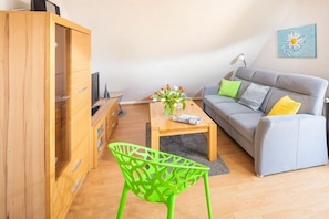 Ein Wohnzimmer mit grauem Sofa, einem Holzkasten, einem grünen Sessel und einem Couchtisch mit Blumen.