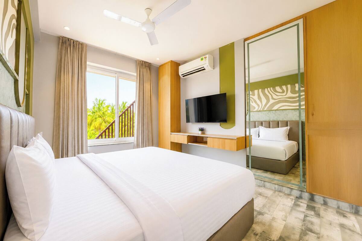 Habitación de hotel con cama, televisión, aire acondicionado y balcón con vistas a palmeras.