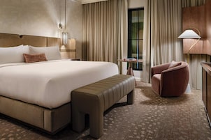 Habitación de hotel con una cama grande, un banco, una silla, una mesita de noche y una lámpara.