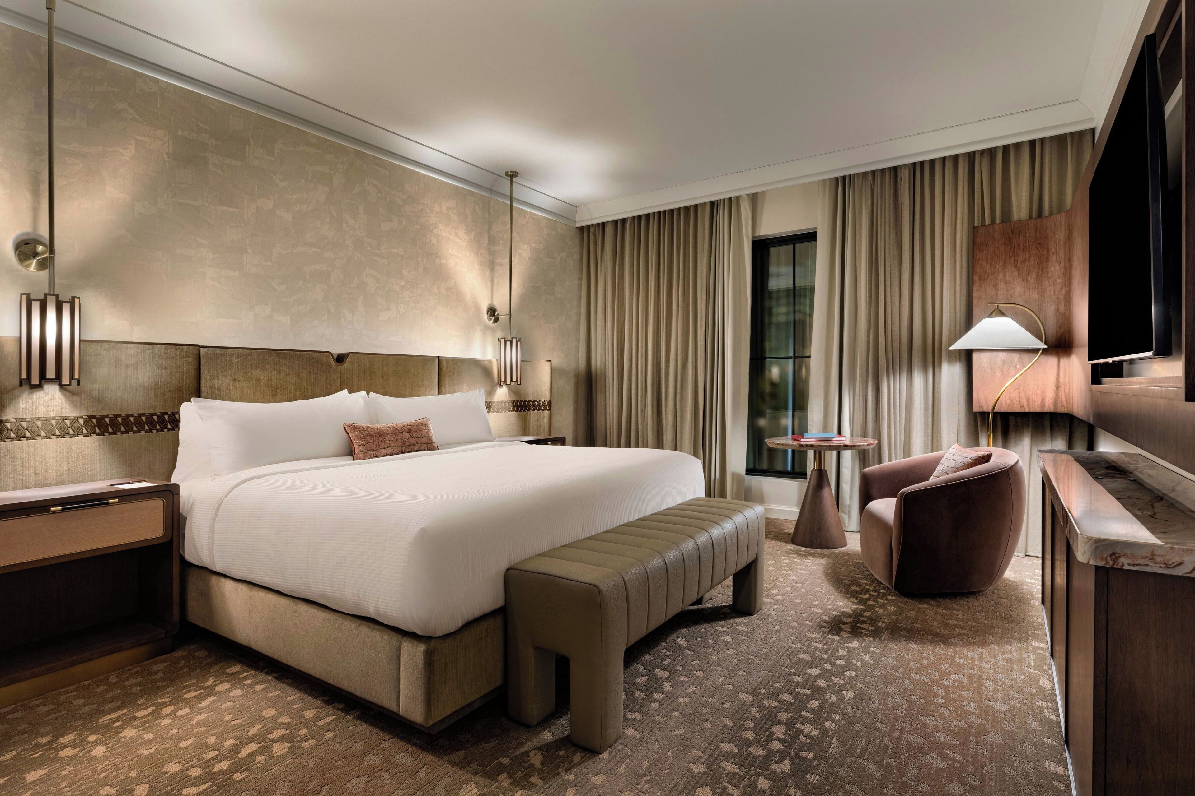 A hotel room with a large bed, a bench, a desk, a chair, a TV, and a small table.