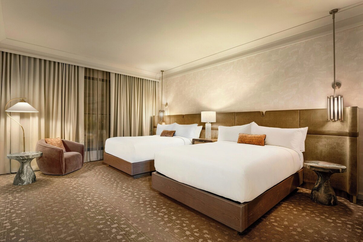 Habitación de hotel con dos camas, una zona de estar y un ventanal grande con cortinas.