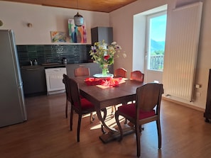 Un coin repas avec une table et des chaises en bois, un vase de fleurs et, au fond, une cuisine.