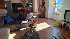 Un salon moderne avec un canapé, une table basse ornée d’un vase de fleurs, un téléviseur et une grande fenêtre donnant sur un beau paysage.