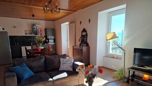 Un salon avec un canapé, une table basse ornée d’un vase de fleurs, un téléviseur, et un coin cuisine à l’arrière-plan.