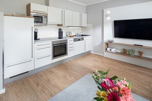 A modern kitchen with white cabinetry, a built-in oven, microwave, and a large flat-screen TV mounted above a wooden shelf with decorative items. - Düsseldorfer Hof A1 - first class living (Norderney)