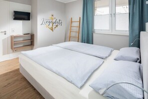 A modern hotel room with a bed, a mounted TV, a wooden ladder, and a window with curtains. - Düsseldorfer Hof A1 - first class living (Norderney)