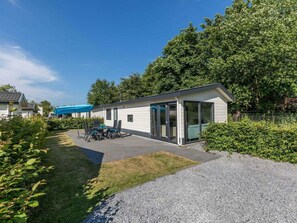 Ein modernes Mobilheim mit überdachter Terrasse, kleinem Garten und gepflasterter Auffahrt.
