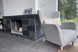 Ein modernes Wohnzimmer mit Kamin, einem grauen Sessel und Blick auf Bäume durch ein gro ßes Fenster.