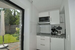 Eine kleine Küche mit weißen Schränken, einer Mikrowelle, einer Kaffeemaschine und Blick auf einen Garten durch eine Glastür.
