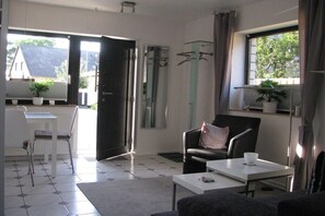 Ein Zimmer mit einem weißen Tisch, einem schwarzen Sessel, einer Couch, einem Fenster mit Blick auf Bäume und einer Tür zum Ausgang.