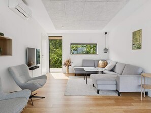 Un salón moderno con un sofá y un sillón grises, un televisor de pantalla plana y un ventanal con vistas a zonas verdes.