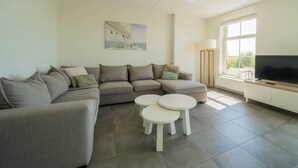A modern living room with a sectional sofa, two small round tables, a flat-screen TV, and a large window. - Westertoren Sauna 12 (Uitdam)