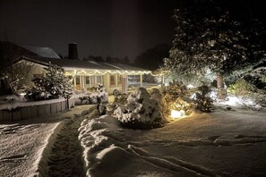 Un paysage enneigé avec une maison illuminée par des lumières, entourée d’arbres chargés de neige.