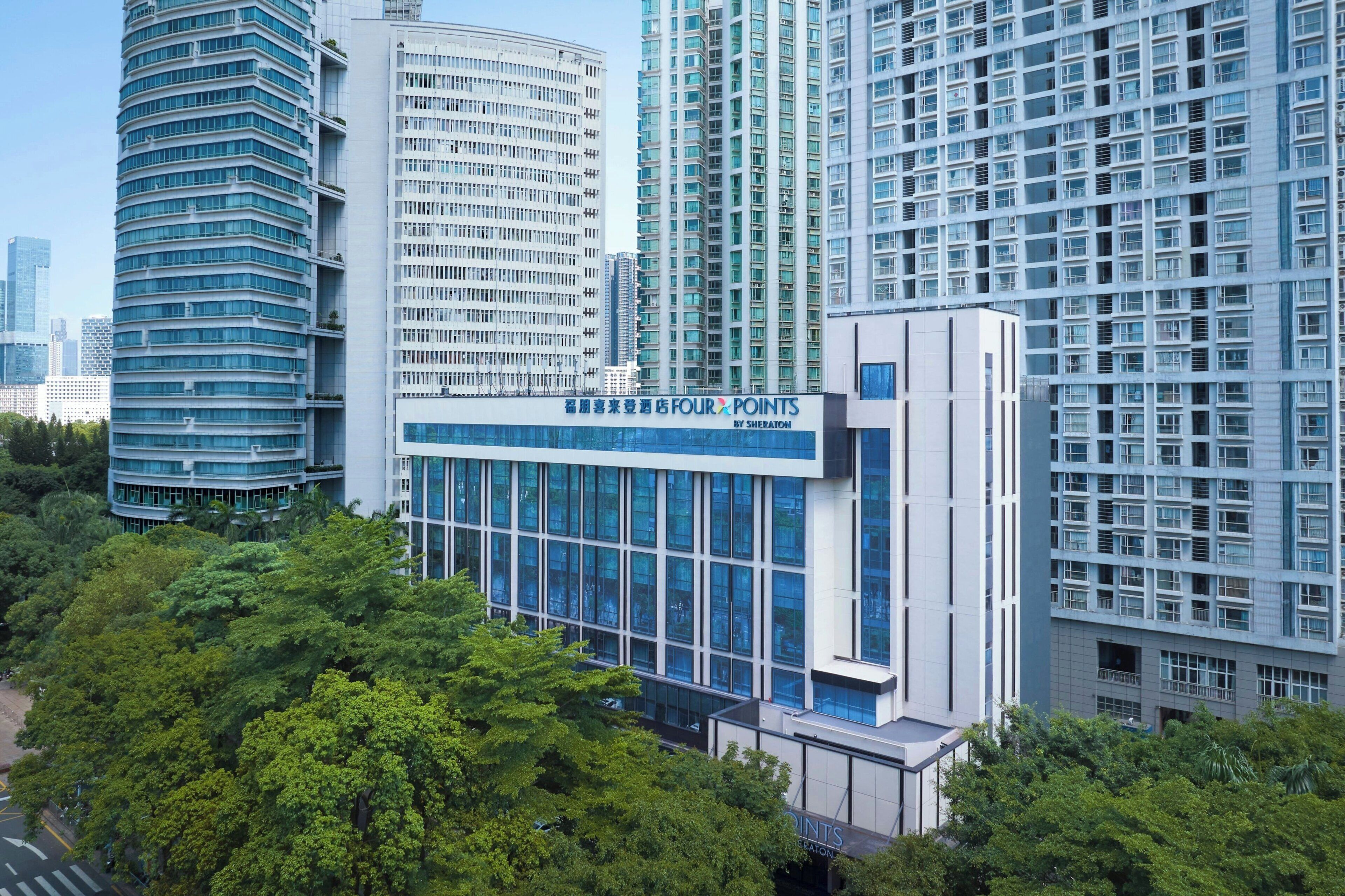 Un moderno edificio hotelero con fachada de vidrio, rodeado de rascacielos y abundante vegetación.