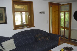Ein Zimmer mit einem blauen, gemusterten Sofa, einem weißen Kissen, einem kleinen Tisch mit Teller und Kerze, einem Fenster mit Vorhang und einer Tür mit Glasfeldern.