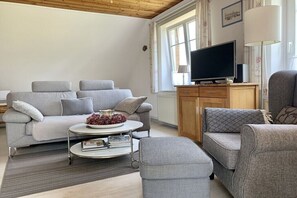 A living room with a sofa set, a coffee table with books and a bowl of grapes, a television on a stand, and a lamp. - Holiday apartment with 2 bedrooms (Tönning)