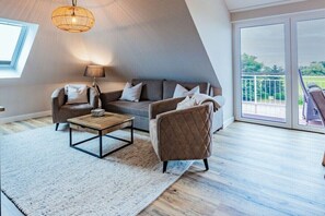 Ein modernes Wohnzimmer mit Sofa, Ohrensessel, Couchtisch und einem großen Fenster mit Blick ins Freie.