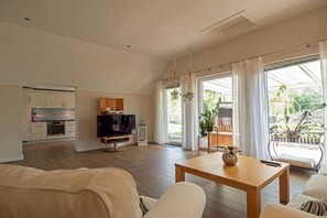Ein modernes Wohnzimmer mit Couch, Fernseher und einem Holztisch. Es gibt eine Küchenzeile mit Schränken und einem Kühlschrank. Große Fenster sorgen für natürliches Licht und einen Blick ins Freie.