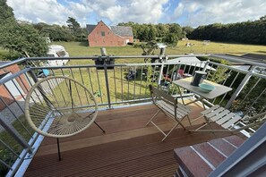 Eine Holzterrasse mit einer Hängematte, einem Tisch und Stühlen mit Blick auf ein Wiesfeld und einen roten Bauernhof.