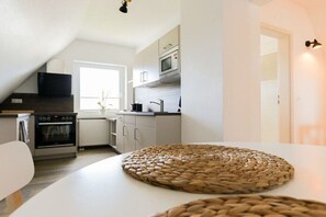 A modern kitchen with white cabinetry, a microwave, an oven, and a large woven placemat on the table. - 2 Zeitke "the Little One" (Altefähr)