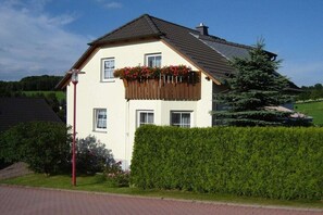 A two-story house with a sloped roof, a wooden balcony with flowers, a red street lamp, and a hedge in the foreground. - in the Bergblick house (Annaberg-Buchholz OT Cunersdorf)