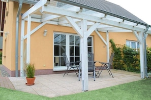 Eine überdachte Terrasse mit Tisch und Stühlen, einem Blumentopf und einem Haus mit gelbem Mauerwerk.