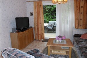 Ein Wohnzimmer mit Fernseher auf einer Holzkommode, einer Couch, einem Couchtisch mit Blumen und Blick ins Freie durch eine Glastür.