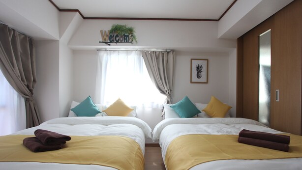 Habitació d'hotel amb dos llits, cadascun amb una manta groga i una tovallola marró, roba de llit blanca i un cartell amb la paraula 'Welcome' sobre la finestra.