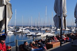 Ein Yachthafen mit vertäuten Booten, Personen, die auf einer Terrasse sitzen, und Sonnenschirmen, die Schatten spenden.