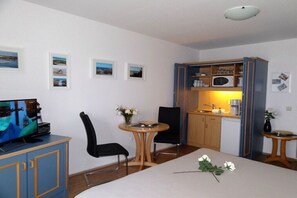 Ein Zimmer mit einem Bett, einem Schreibtisch, einem Sessel, einem Fernseher, einem kleinen Tisch mit Blumen und einer Küchenzeile.