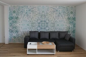 Ein Wohnzimmer mit ornamental gestalteter Tapete, einem schwarzen Sofa mit Kissen und einem Holztisch.