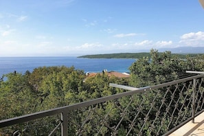 Ein Balkon mit Metallgeländer, der auf eine Küstenlandschaft mit Bäumen und eine entfernte Meer- und Hügellandschaft blickt.