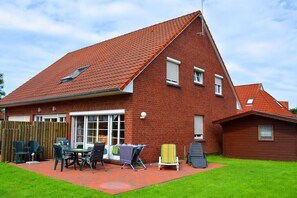 Ein rotes Backsteinhaus mit geneigtem Dach, einer Terrasse und Gartenmöbeln sowie einem Holzzaun.