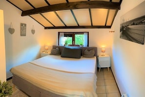 Ein Schlafzimmer mit einem großen Bett, zwei Nachttischen mit Lampen, einem Fenster mit Vorhängen und einer Decke mit freiliegenden Balken.
