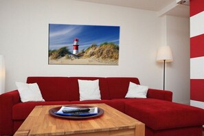 A red sofa with white cushions, a wooden coffee table with a remote control, and a framed picture of a lighthouse on the wall. - Holiday apartment with 1 bedroom (Borkum)