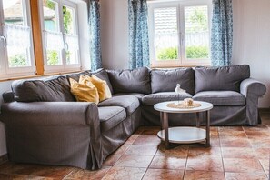 Ein graues Ecksofa mit einem kleinen runden Tisch in einem Raum mit Fliesenboden und großen Fenstern mit Vorhängen.
