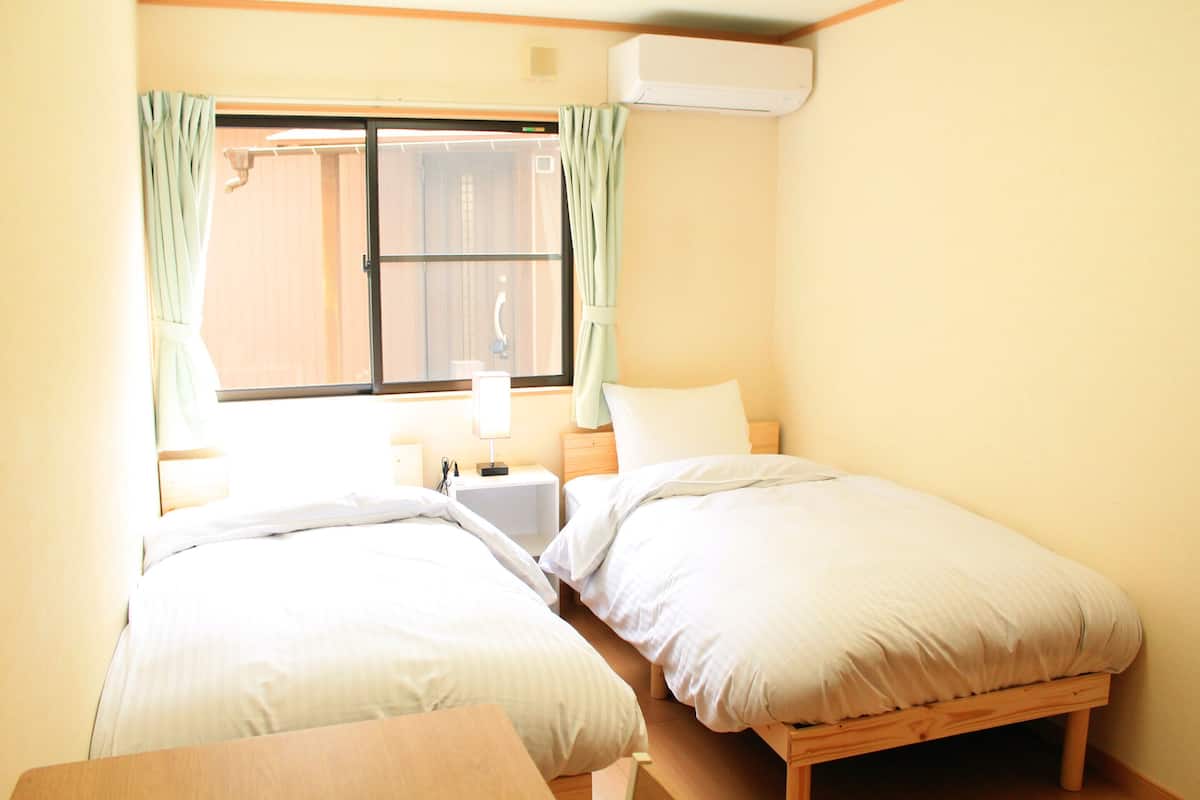 A small, well-lit room with two beds, a window with curtains, a bedside table with a lamp, and an air conditioning unit.