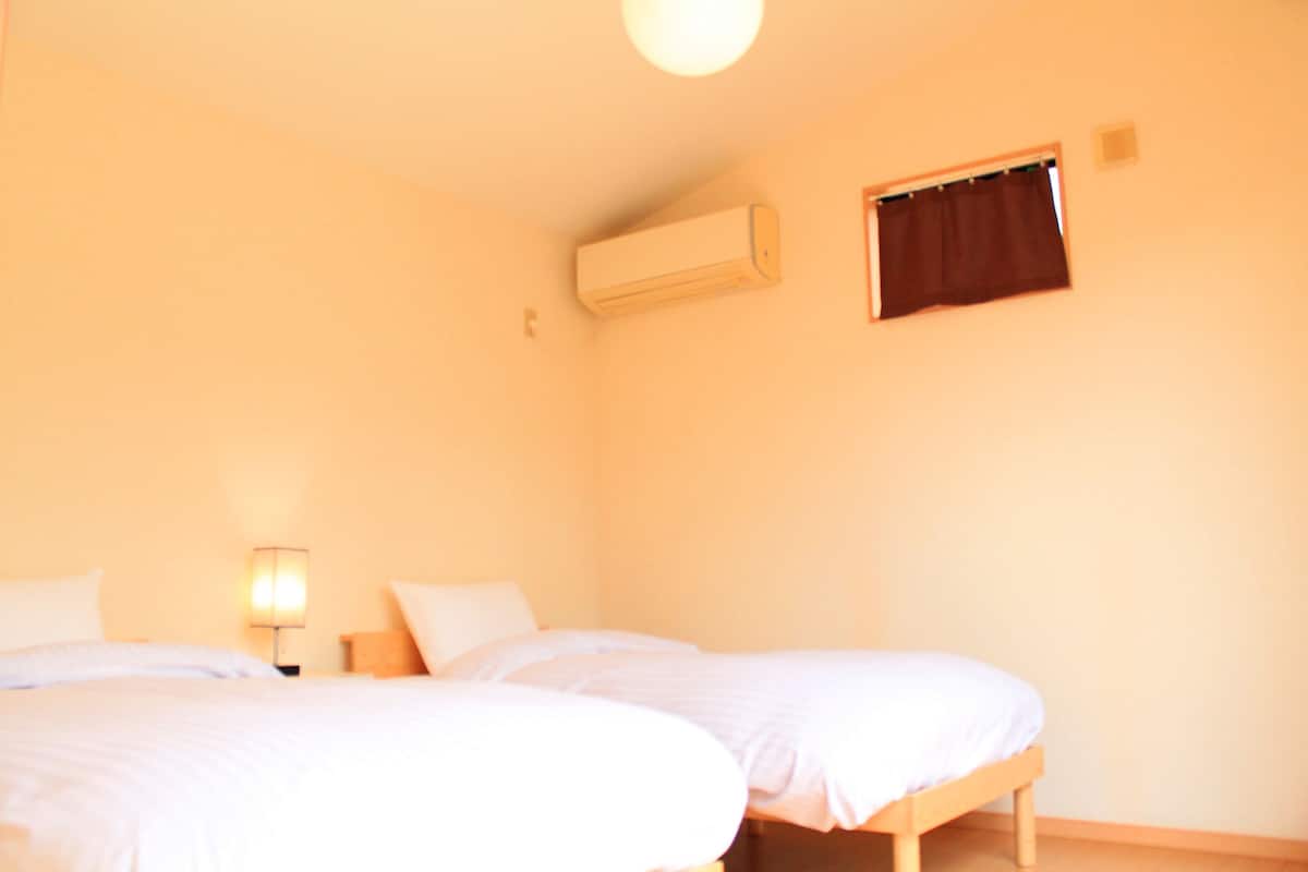 A room with two beds, a wall-mounted air conditioner, a small window with a curtain, and a bedside lamp.