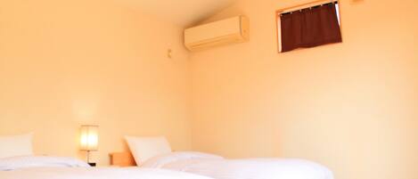 A room with two beds, a wall-mounted air conditioner, a small window with a curtain, and a bedside lamp.