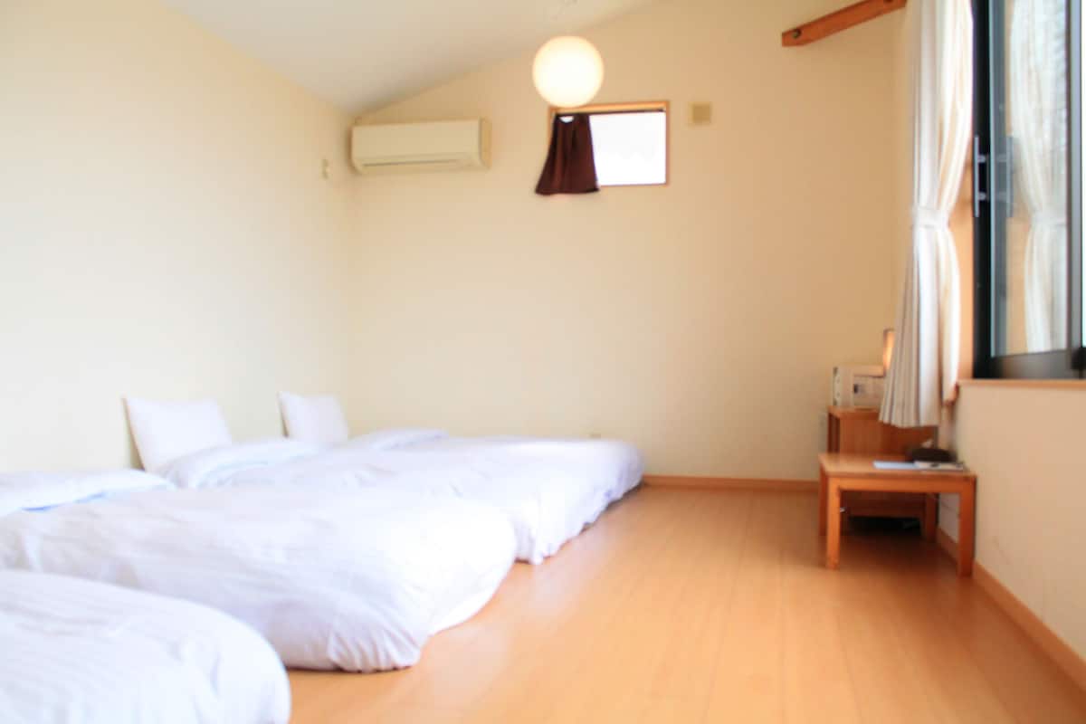 A room with two beds, a small table, an air conditioning unit, and a window with curtains.
