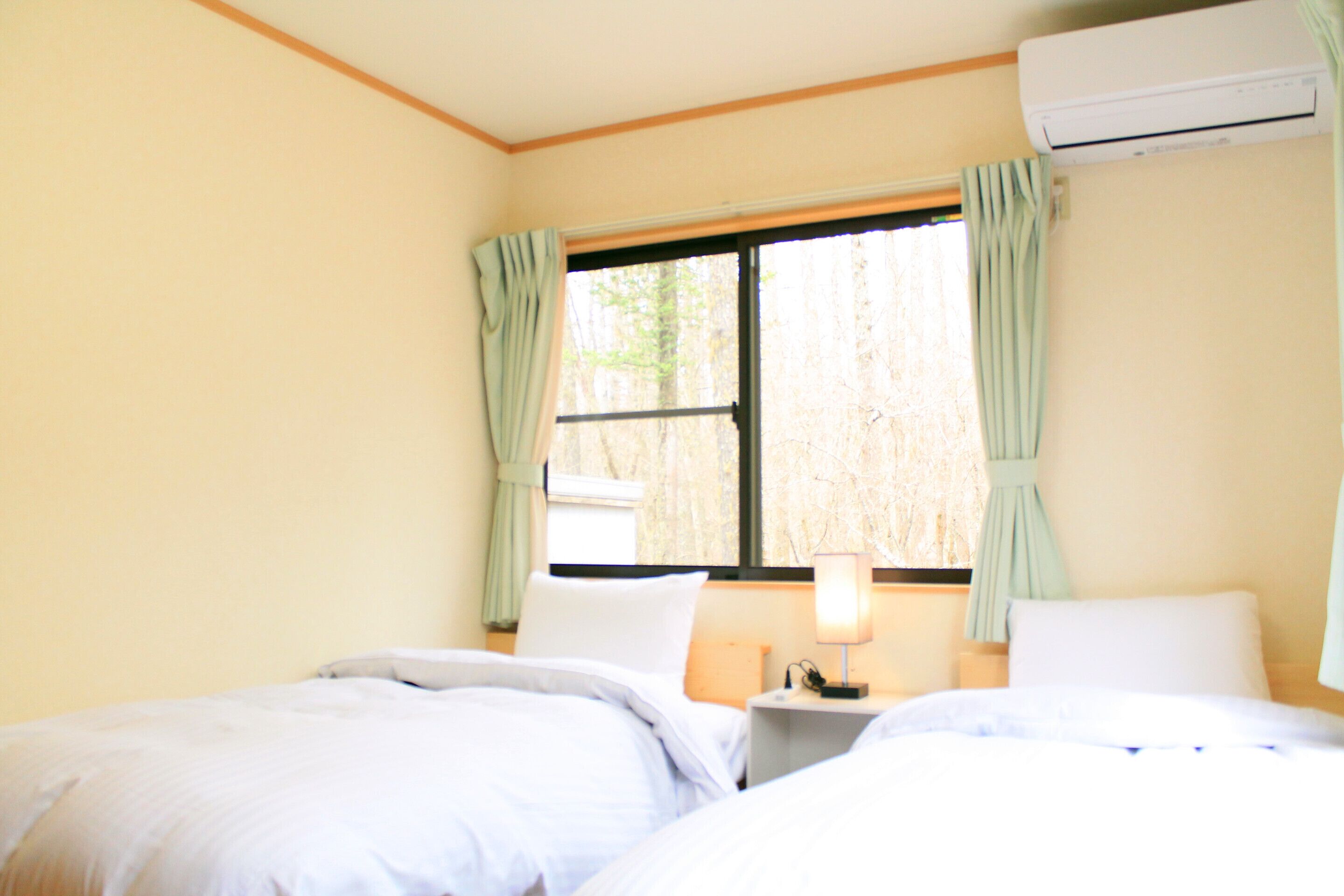 Una habitación con dos camas, una mesita de noche con lámpara, aire acondicionado y una ventana con cortinas.