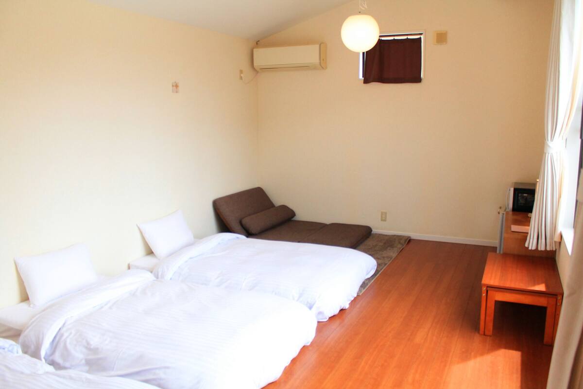 A room with two beds, a brown chair, a small wooden table, an air conditioning unit, and a window with curtains.