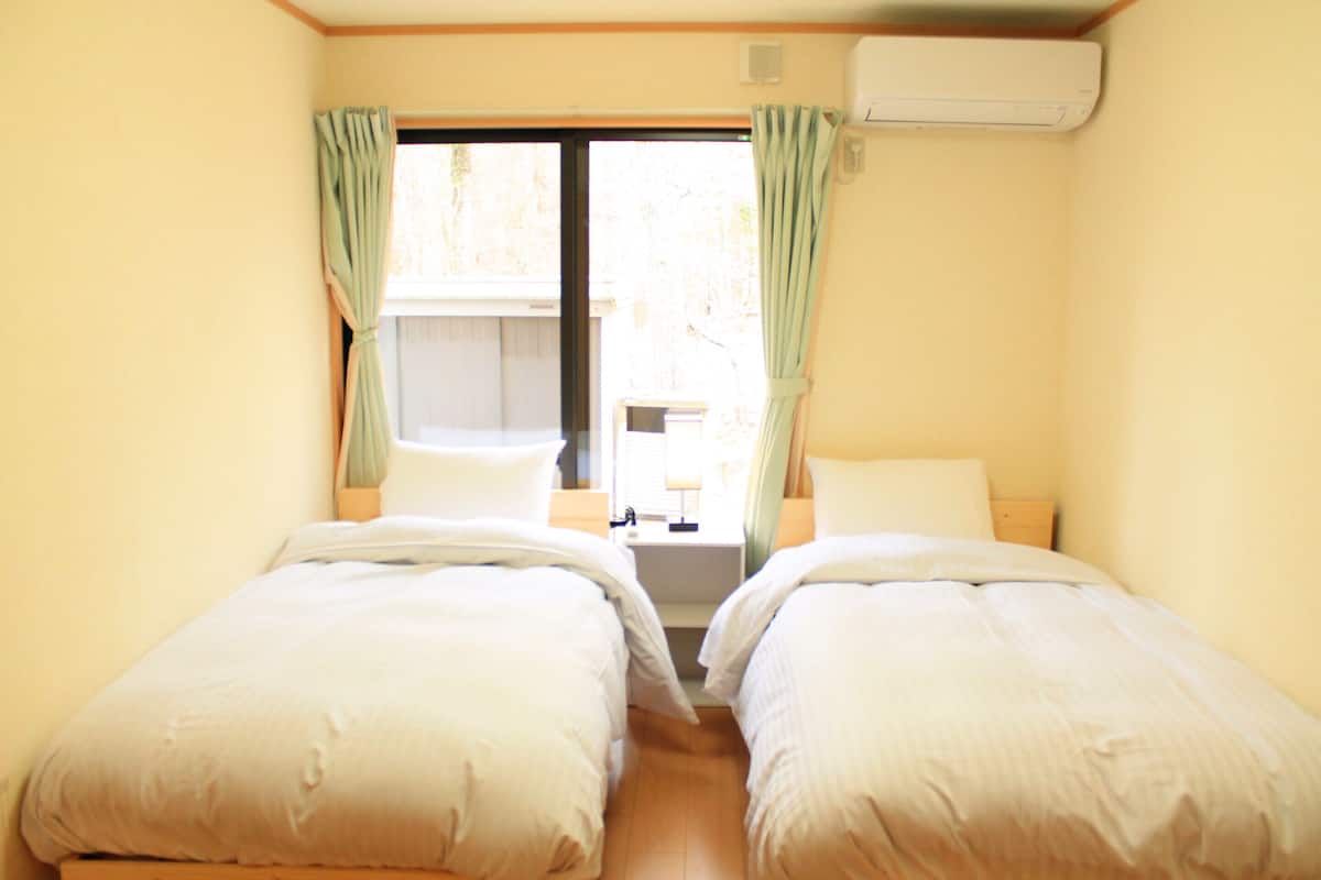 A room with two beds, a window with curtains, a bedside table, and an air conditioning unit.