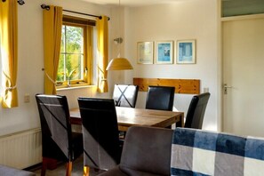 A dining area with a wooden table, black chairs, and yellow curtains. There are framed pictures on the wall and a window with curtains. - Holiday home Strandperle 174 (Julianadorp)